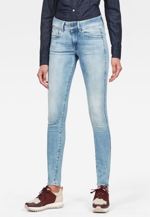 Persoon draagt lichtblauwe skinny jeans, donkerblauw shirt met lange mouwen en beige-bruin sneakers, staand met ontspannen handen.