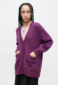 Μωβ πλεκτή ζακέτα με ντεκολτέ σε σχήμα V, μακριά μανίκια και δύο μπροστινές τσέπες. Υφή υφάσματος με ριμπ μανσέτες και κλείσιμο με κουμπιά.