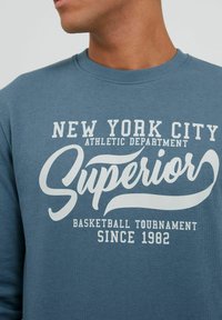 Plavi pamukasti dukserica s bijelim tekstom: "ATLETSKI ODJEL GRADA NEW YORKA," "Superior," i "TURNIR U KOŠARCI OD 1982."