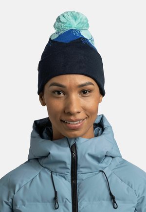 Gorro de lana con una base azul oscura, que presenta un pom-pom azul claro y patrones de montañas. Lleva puesto un abrigo azul claro, que muestra cremalleras.