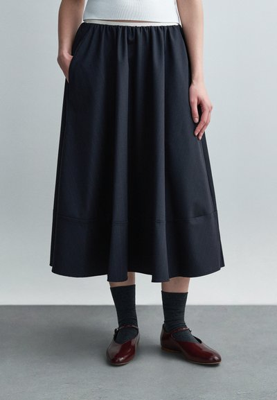 Personne portant une jupe midi noire, des chaussettes foncées et des chaussures Mary Jane bordeaux brillantes, debout sur un sol gris devant un fond clair.