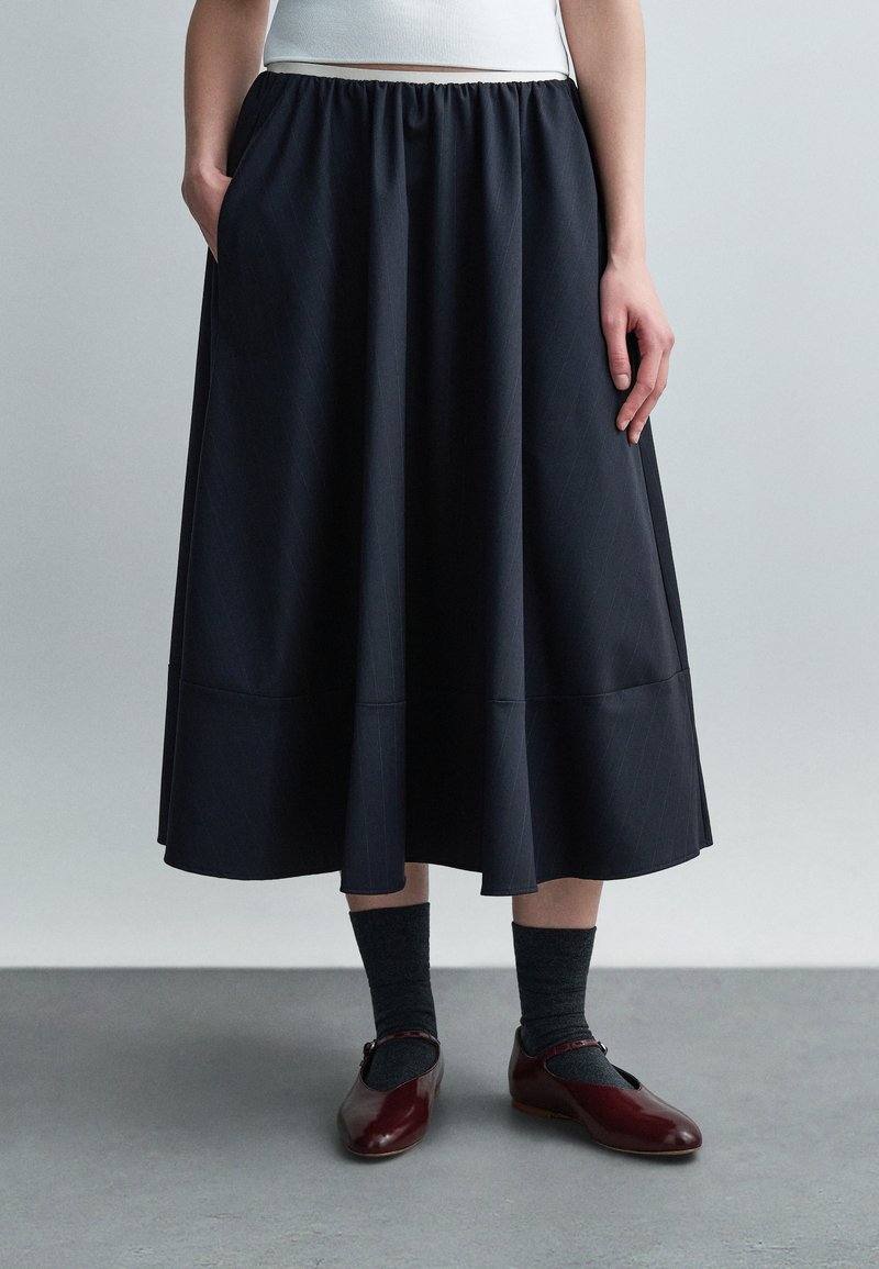 Άτομο που φοράει μαύρη midi φούστα, σκουρόχρωμες κάλτσες και λαμπερές μπορντό γόβες Mary Jane, στέκεται σε γκρι πάτωμα με φωτεινό φόντο.