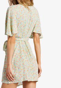 Blumenkleid in Hellgrün mit rosa und orangen Blumen. Verfügt über kurze, fließende Ärmel und eine gebundene Taillierung. Strukturierten Stoff.