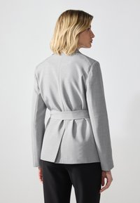 Veste grise au design structuré, taille ceinturée et détails plissés à l'arrière. Le tissu semble lisse avec une coupe ajustée.