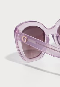 Gafas de sol moradas con un diseño geométrico estilo gato, con un marco translúcido y lentes oscuros degradados. Logo dorado de la "G" en la patilla.