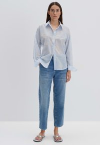 Camicia azzurro chiaro in tessuto trasparente, abbinata a jeans in denim blu sbiadito con cuciture in evidenza e tasche applicate. Sandali argentati.