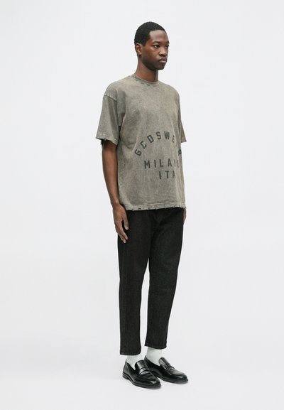 Tricou supradimensionat de culoare verde măslin estompat, cu text negru, asociat cu pantaloni negri conici și loafers negri strălucitori, purtat cu șosete albe.
