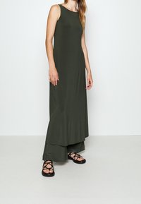 Dlouhé olivově zelené šaty vyrobené z hladké, lehké tkaniny, bez rukávů a s vrstveným spodním lemem; doplněné o černé sandály na řemínky.