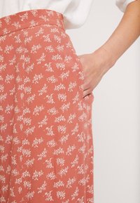 Des pantalons en tissu couleur rouille à motif floral présentent une taille haute et des poches latérales. Le matériau doux est léger avec une texture lisse.