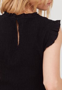 Donna con capelli biondi che indossa una blusa senza maniche nera con bordi arricciati e un'apertura a chiave sul retro del collo.