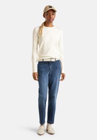 Hellebeige Pullover, dunkelblaue Jeans, weißer Gürtel, beigefarbene Mütze, cremefarbene Loafers und weiße Crew-Socken, stehend vor einem neutralen Hintergrund.