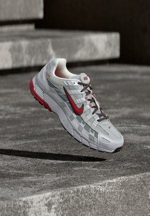 Bielo-sivá bežecká topánka Nike s červenými detailmi a čiernymi šnúrkami, visiaca nad sivými betónovými schodmi.