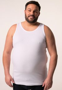 Bílý žebrovaný tílko s kulatým výstřihem a širokými průramky, které nosí muž s vousy na neutrálním pozadí.