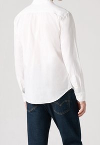 Homme portant une chemise blanche à manches longues boutonnée et un jean bleu foncé, vu de dos sur un fond uni.