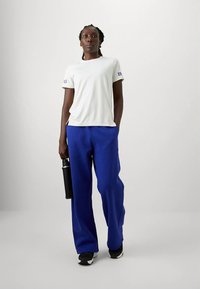 Pessoa de pé com calças azuis e camisa de mangas curtas brancas, segurando uma garrafa de água preta, contra um fundo branco liso.