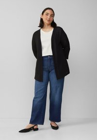 Zwarte cardigan met een open voorkant, gedragen over een wit shirt en blauwe wijde jeans. Zwarte platte schoenen maken de outfit compleet. Glad stoftextuur.