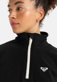 Black fleece pullover with high collar, half-zip front, and small white heart logo on the left chest. Soft texture and simple design.