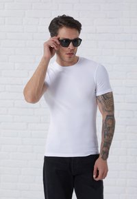 Weißes, kurzärmeliges, figurbetontes T-Shirt mit gerippter Textur, kombiniert mit eleganten schwarzen Sonnenbrillen. Das Modell hat ein kunstvolles Tattoo am rechten Arm.