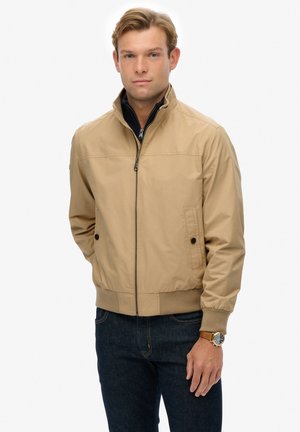Lichtbeige bomberjack met een hoge kraag, rits aan de voorkant, twee zijzakken en elastische manchetten. Draagt over een donker shirt en blauwe spijkerbroek.