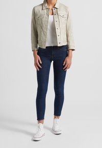 Giacca di jeans grigia chiara con design corto, due tasche sul petto e bottoni argentati, abbinata a jeans skinny blu scuro e sneakers bianche.