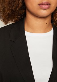 Blazer negro con solapa estructurada y textura suave, superpuesto a una camisa blanca básica. Primer plano del área del cuello y los hombros.