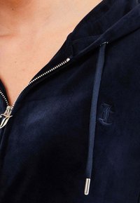 Felpa con zip di velluto blu navy, dotata di una cerniera argentata, cappuccio regolabile con cordino e logo in rilievo. Tessuto morbido con un design aderente.