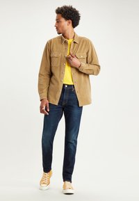 Hombre que lleva una chaqueta de pana color crema sobre una camiseta amarilla, jeans azul oscuro y zapatillas amarillas, mirando a su izquierda con una mano en la chaqueta.