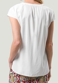Blouse blanche à manches courtes avec un détail froncé au dos, présentant un tissu léger et lisse. Associée à un pantalon multicolore à motifs.
