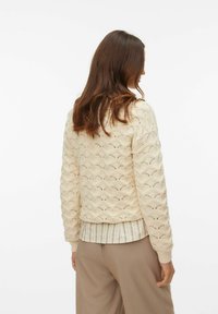 Suéter de punto color crema con un patrón ondulado texturizado, con puños y dobladillo acanalados, superpuesto sobre una camisa a rayas clara.