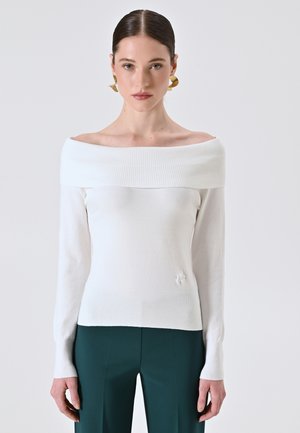 Femme avec des cheveux lisses en arrière, portant des boucles d'oreilles en or, un pull blanc à épaules dénudées et un pantalon vert foncé, se tenant devant un fond uni.