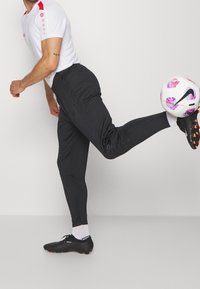 Man i svarta byxor och vit sporttröja som sparkar en vit och lila Nike fotboll mot en enkel vit bakgrund.