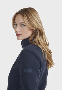 Veste en polaire marine avec un col haut, tissu texturé et un logo AIGLE sur la manche gauche, dotée d'une coupe ajustée.