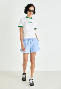 Weißes T-Shirt mit grünem Kragen und Logo, kombiniert mit hellblauen Shorts und schwarzen Sneakers mit weißen Akzenten. Modell trägt dunkle Sonnenbrillen.