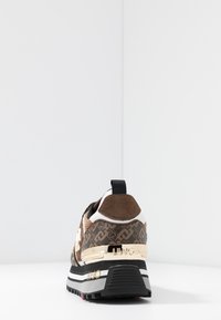 Zapatilla marrón con material estampado, detalles dorados, suela negra y detalles en blanco; cuenta con un talón texturizado y el logo de la marca.