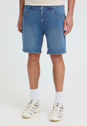 Homme portant un short en jean bleu mi-long, des chaussettes blanches montantes et des chaussures de sport blanches, debout devant un fond uni.