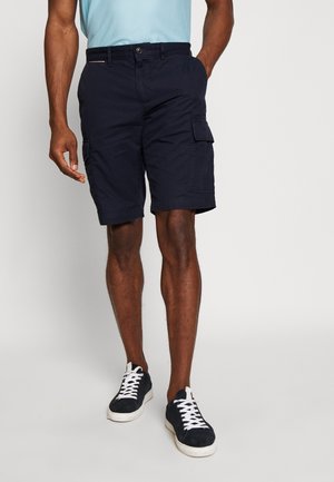 Marineblaue Cargo-Shorts mit Knopfverschluss, Klappentaschen an den Seiten und umgeschlagenem Saum. Getragen mit navyfarbenen Sneakers mit weißen Schnürsenkeln.