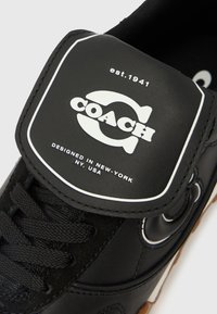 Schwarzer Sneaker mit glattem Lederobermaterial, strukturierten Schnürsenkeln, Gummisohle und einem weißen Logo-Patch mit der Aufschrift "COACH" und "Designed in New York."