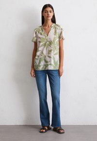 Blouse florale, vert clair et beige à manches courtes avec un décolleté en V et des détails de boutons, assortie à un jean bleu et des sandales noires.