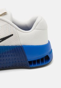 Baskets blanches de sport avec tige en mesh, logo noir et talon en caoutchouc bleu. Présente des détails texturés et une découpe de ventilation sur la semelle.