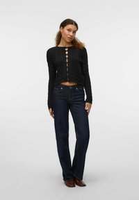 Črni, kratki pleteni pulover z teksturiranimi vzorci in zapiranjem na gumb, v kombinaciji z temno modrimi ravnimi jeans hlačami in rjavimi čevlji.