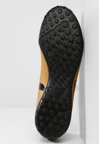 Mustan ja tan värin urheilujalkineen ulkopohja, jossa on teksturoitu kumipinta, jossa on ympyrämuotoisia tartuntoja ja Adidas-logo.