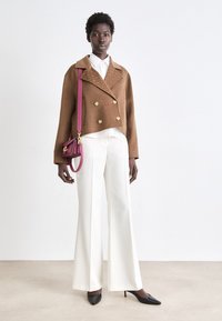 Brown double-breasted wool jacket with gold buttons, white blouse, and wide-leg white trousers. Black pointed heels and a pink crossbody bag.