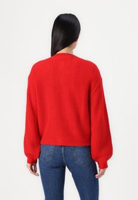 Femme aux longs cheveux noirs portant un pull en maille rouge vif et un jean bleu, debout de dos sur un fond blanc uni.