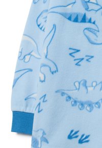 Manta de felpa azul claro con patrones de dinosaurios de dibujos animados y aviones en un azul más oscuro. Textura suave con detalles de puño elástico.