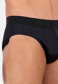 Pantaloni slip neri da uomo con una texture morbida e liscia e una cintura elastica larga con branding in rilievo. Taglio alto sulla coscia.