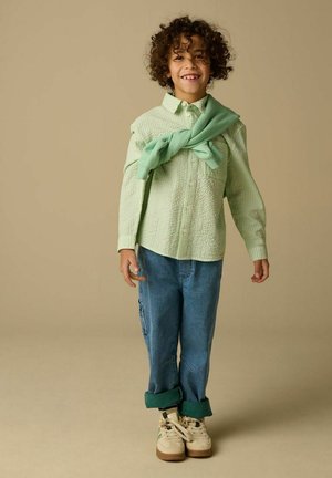 Jeune enfant aux cheveux bouclés debout, portant une chemise texturée vert clair, un jean bleu retroussé, des baskets beiges et un pull vert posé sur les épaules.