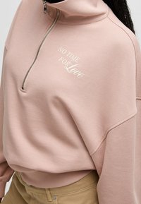 Jasnoróżowa bluza z wysokim kołnierzem, zamkiem na pół długości i białym napisem "NO TIME FOR Love." Miękki materiał z prążkowanymi mankietami i dołem.