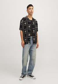 Camisa negra de manga corta con un patrón de hojas de palma blancas, combinada con unos pantalones vaqueros holgados de color azul claro y zapatillas negras.