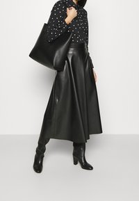 Czarna skórzana spódnica o wysokim stanie i pełnym kształcie, zestawiona z czarną bluzką w białe kropki oraz czarną torbą na ramię.