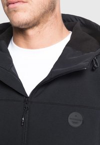 Giacca nera con zip e cappuccio, caratterizzata da una texture liscia e un patch con logo discreto sulla parte sinistra del petto. Camicia bianca visibile sotto.